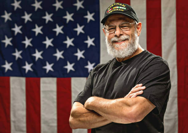 Veteran with white beard and hat in front of American flag.