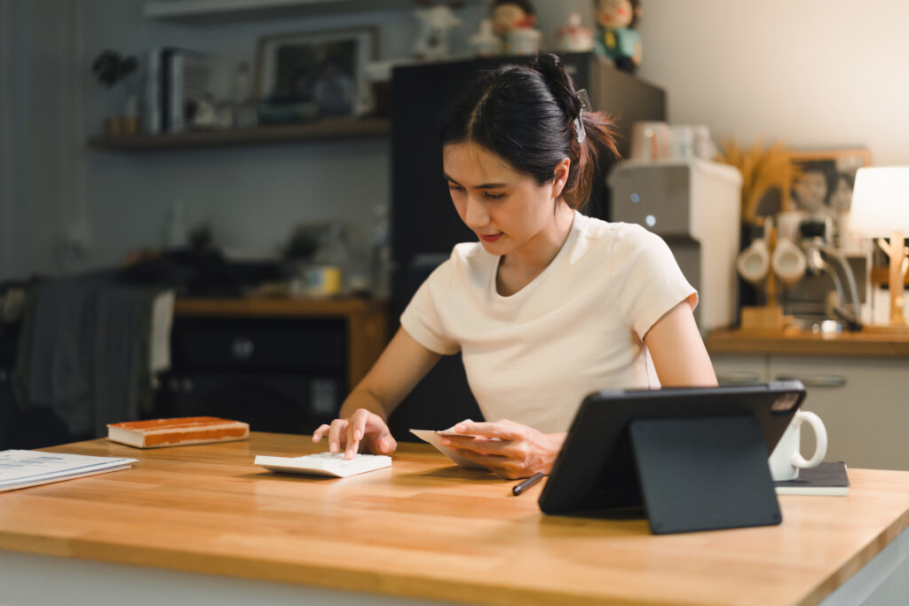 Person using calculator while holding bills reviewing financial documents related to taxes and mortgage. The scene conveys a sense of focus and diligence in managing personal finances.